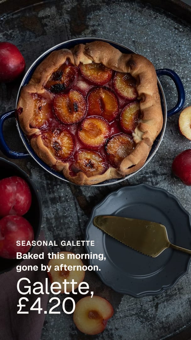 Ad for a coffee shop: a plum galette baked in a dark blue enamel dish, seen from above on a scratched metal tray with halved plums around it. The words on the picture read Seasonal galette, Baked this morning, gone by afternoon, Galette, £4.20.